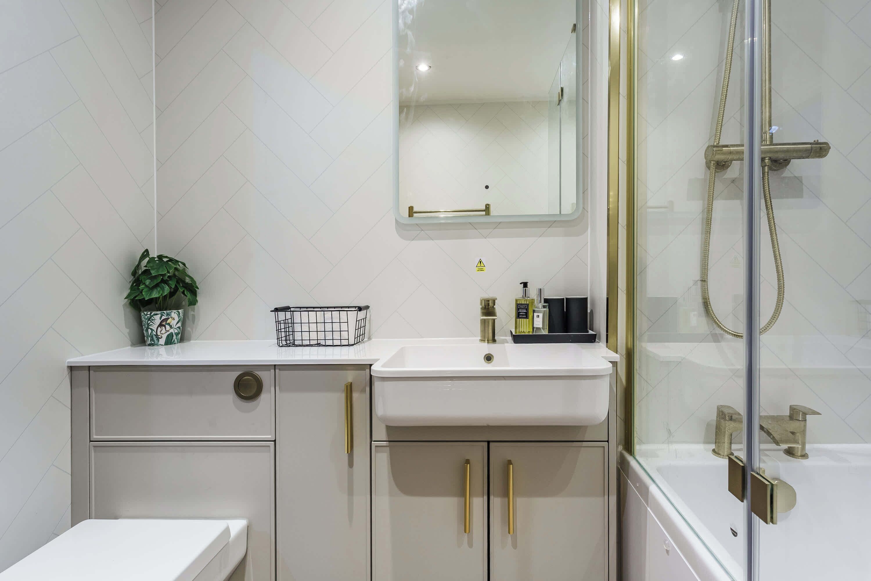 A bathroom with a white sink, gold handles, a mirror, a shower, and a potted plant.
