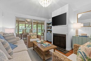 A living room with a sofa set, a coffee table, a fireplace, a television, and a view of the outdoors. - 3BR golf-course cottage on Seabrook Island w/screened porch "Serene Seahorse"-no pets (Seabrook Island)