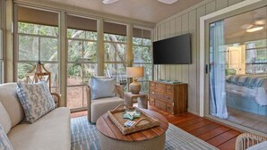 A sunlit room with a ceiling fan, large windows, a flat-screen TV, a sofa, a coffee table, and a wooden cabinet. - 3BR golf-course cottage on Seabrook Island w/screened porch "Serene Seahorse"-no pets (Seabrook Island)