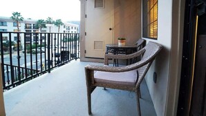 Ein Balkon mit Korbstuhl und kleinem Tisch, eine Tür mit Lüftungsgitter und Blick auf Gebäude und Palmen.