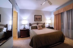 Un dormitorio con una cama, dos mesitas de noche, una lámpara de noche, un ventilador de techo, una puerta y una ventana con cortinas.