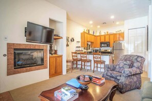 A living room with a fireplace, a television, a dining area with chairs, and a kitchen with wooden cabinets and appliances. - La Quinta Workcation Condo · Fast WiFi & Pool (La Quinta)