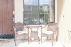 Un patio con sillas de mimbre, una pequeña mesa, una puerta de madera y una ventana con persianas.