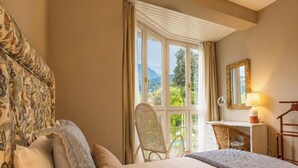 Een slaapkamer met een bed, een stoel, een dressoir, een spiegel en een raam met uitzicht op groen.