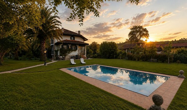 A rectangular swimming pool with a paved border, surrounded by a well-maintained lawn, a palm tree, and a house with a tiled roof. - Rural Villa with Pool in Llanes | La Ablanosa (Llanes)