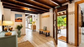 Un passadís amb sòl de fusta, una banca, una làmpada, un vas amb una planta, un quadre penjat a la paret i una porta que dona a zona exterior.
