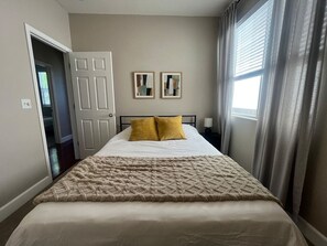 Ein Schlafzimmer mit einem Bett, zwei Kissen, einer Decke, einer Tür, einem Fenster mit Vorhängen und zwei gerahmten Bildern an der Wand.