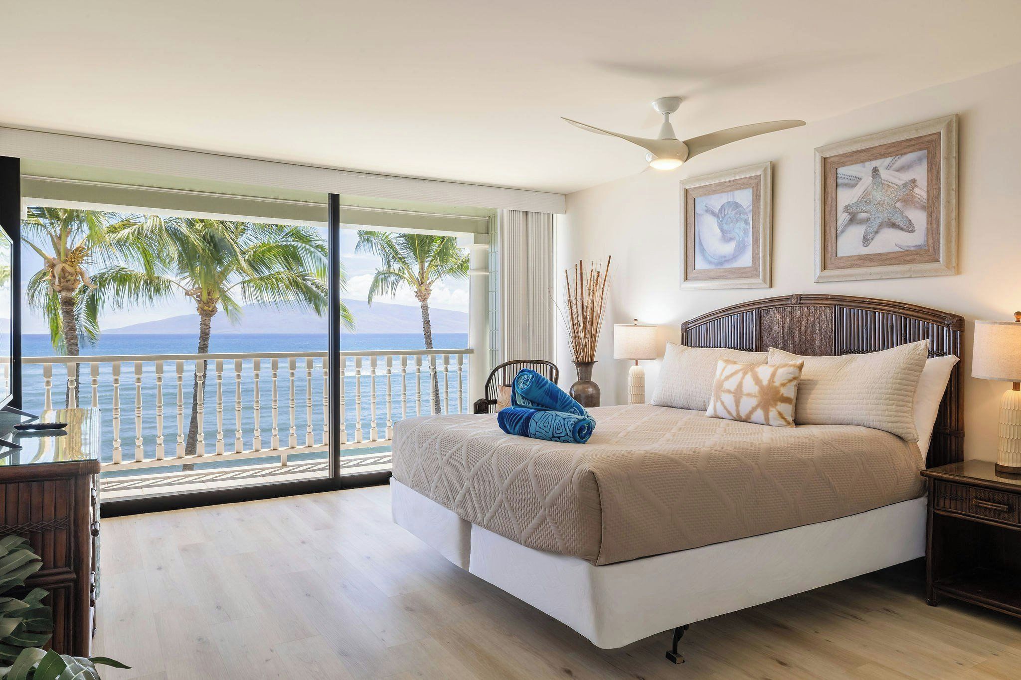 A bedroom with a large bed, a ceiling fan, a nightstand, a lamp, and a view of the ocean through a balcony.