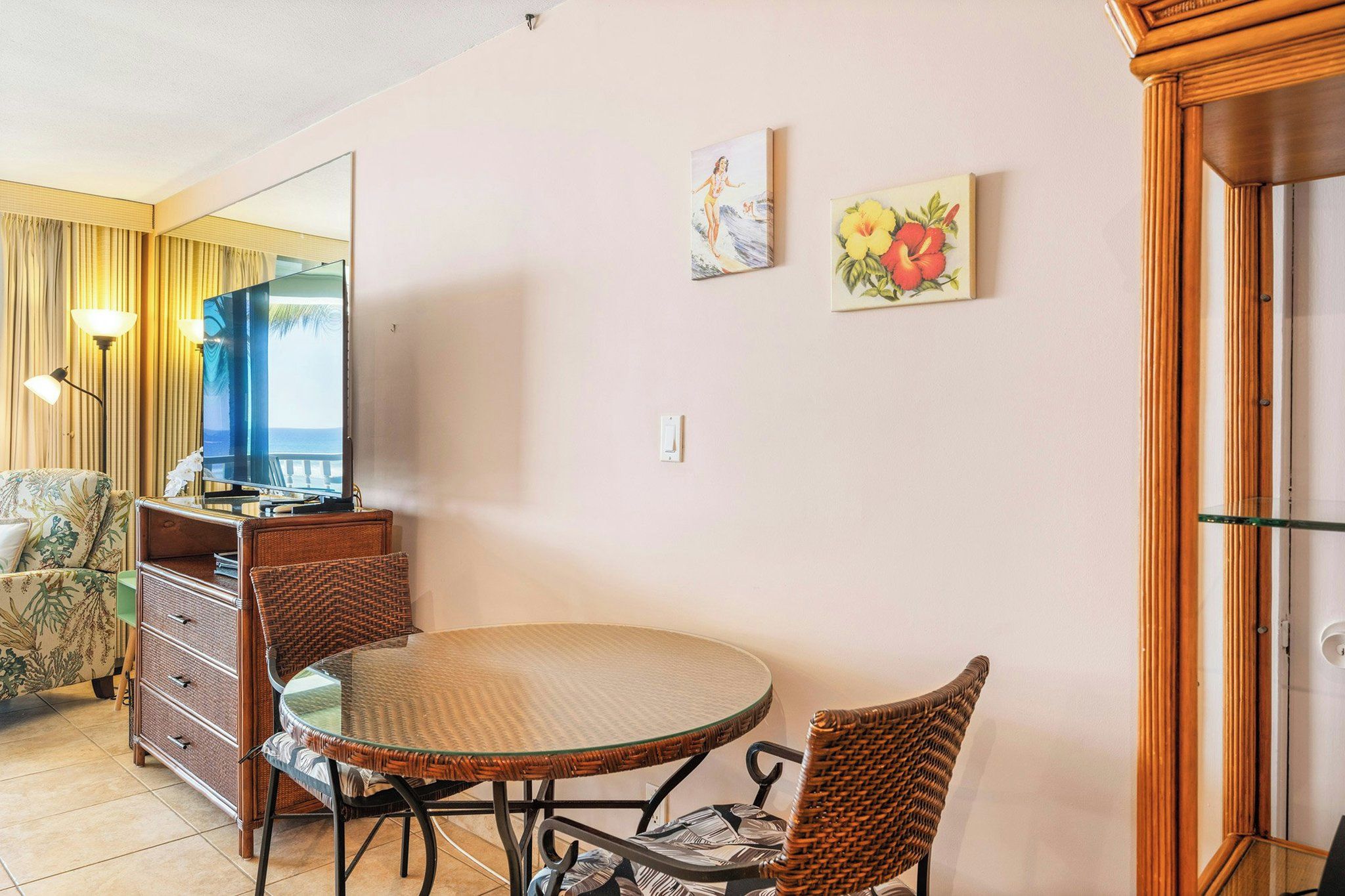 A room with a round glass table, wicker chairs, a dresser, a television, and two wall-mounted paintings.