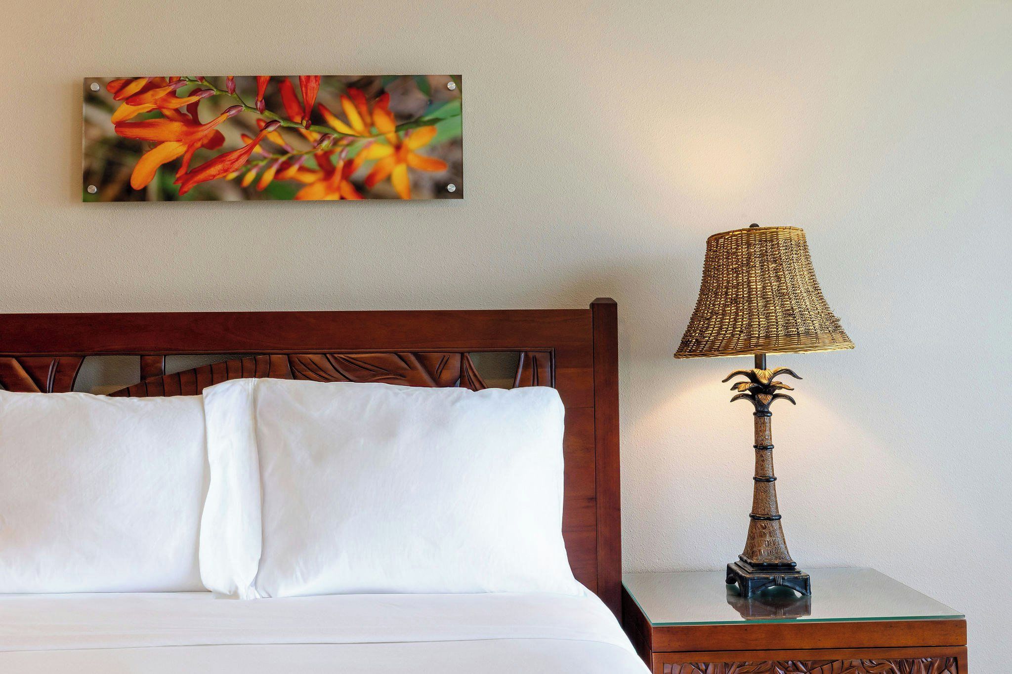 A hotel room with a wooden headboard, a white bed with two pillows, a glass table with a lamp, and a wall-mounted painting.
