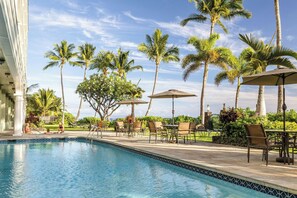 Zona junto a la piscina con vista despejada al océano, rodeada de palmeras y mobiliario exterior.