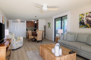 Una sala de estar moderna con sofá, mesa de centro y mueble para televisor. Cuenta con una zona de comedor con sillas y mesa. La habitación tiene un ventilador de techo y vistas al exterior a través de un ventanal.