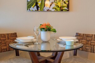 Una mesa redonda de vidrio con platos blancos, un tazón blanco y dos copas de vino, sobre un pared texturizada con diseño de silla tejida.