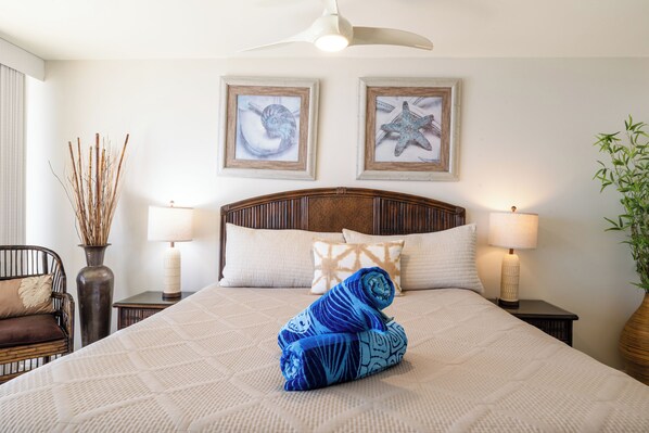 A neatly made bed with a rolled towel, two framed pictures on the wall, a lamp on each bedside table, and a chair with a cushion.