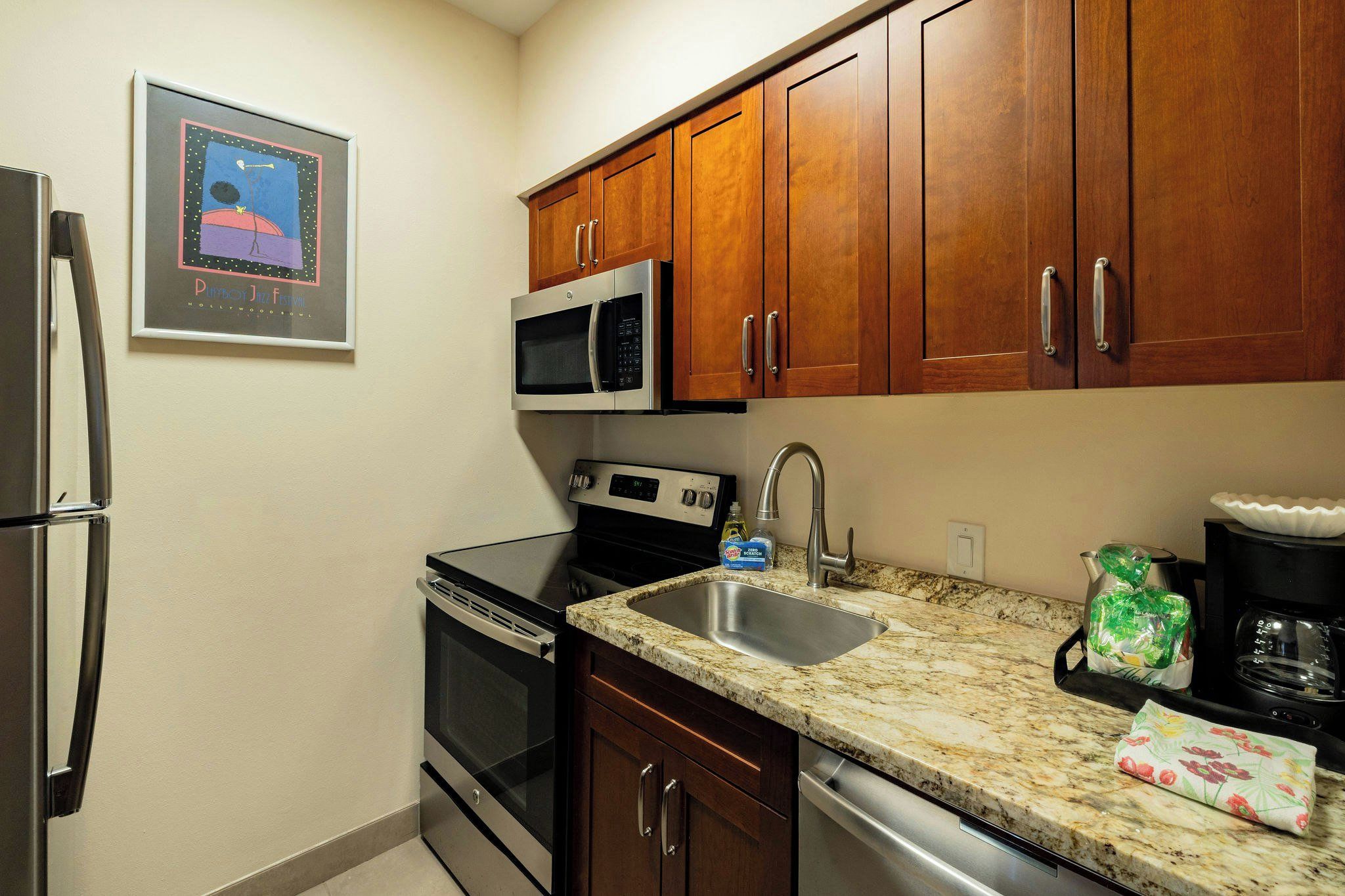 A compact kitchen with a stainless steel refrigerator, a microwave, an oven, a sink, and wooden cabinets.
