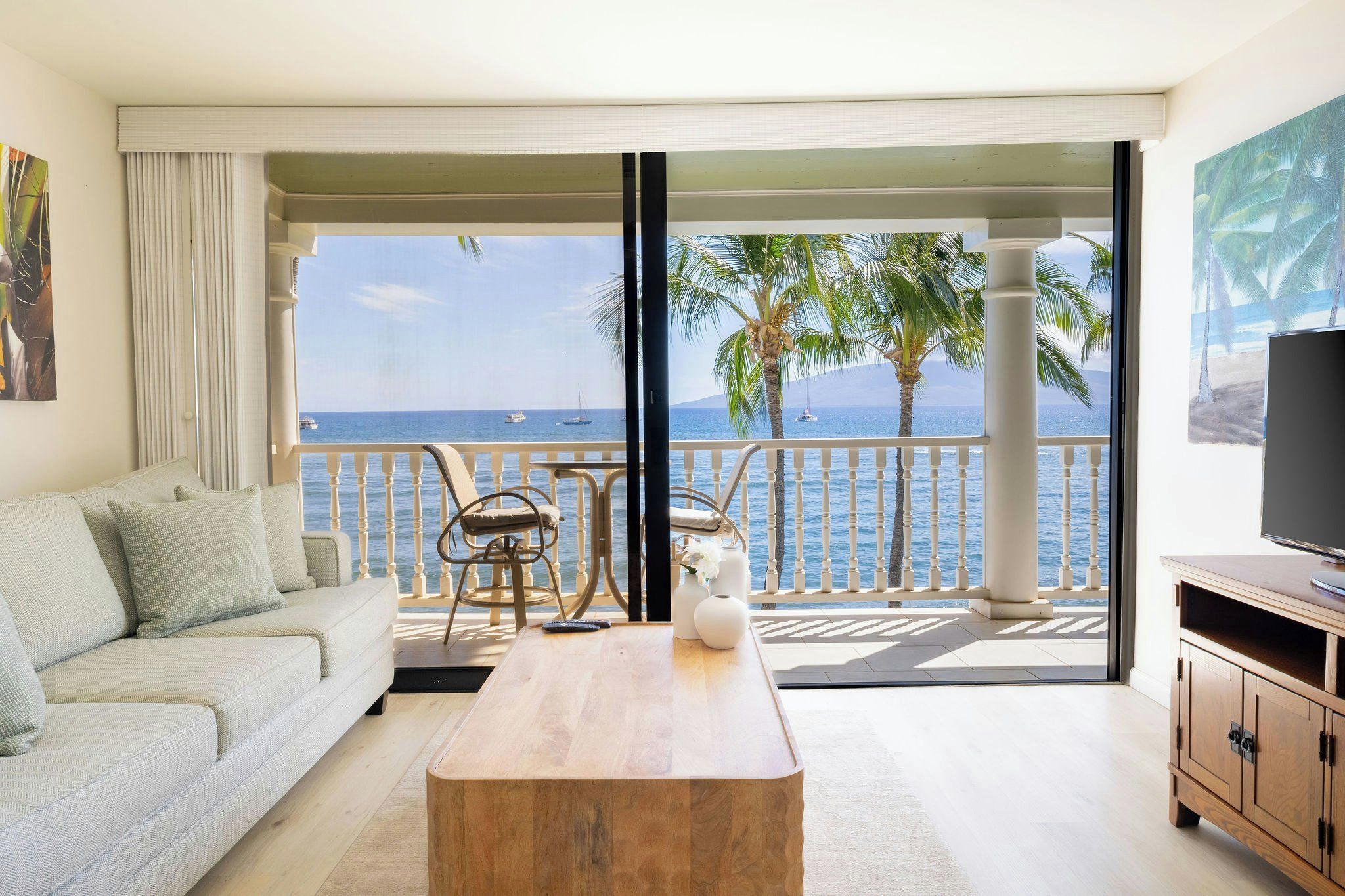 A living room with a sofa, coffee table, and chairs, overlooking a balcony with a view of the sea and palm trees.