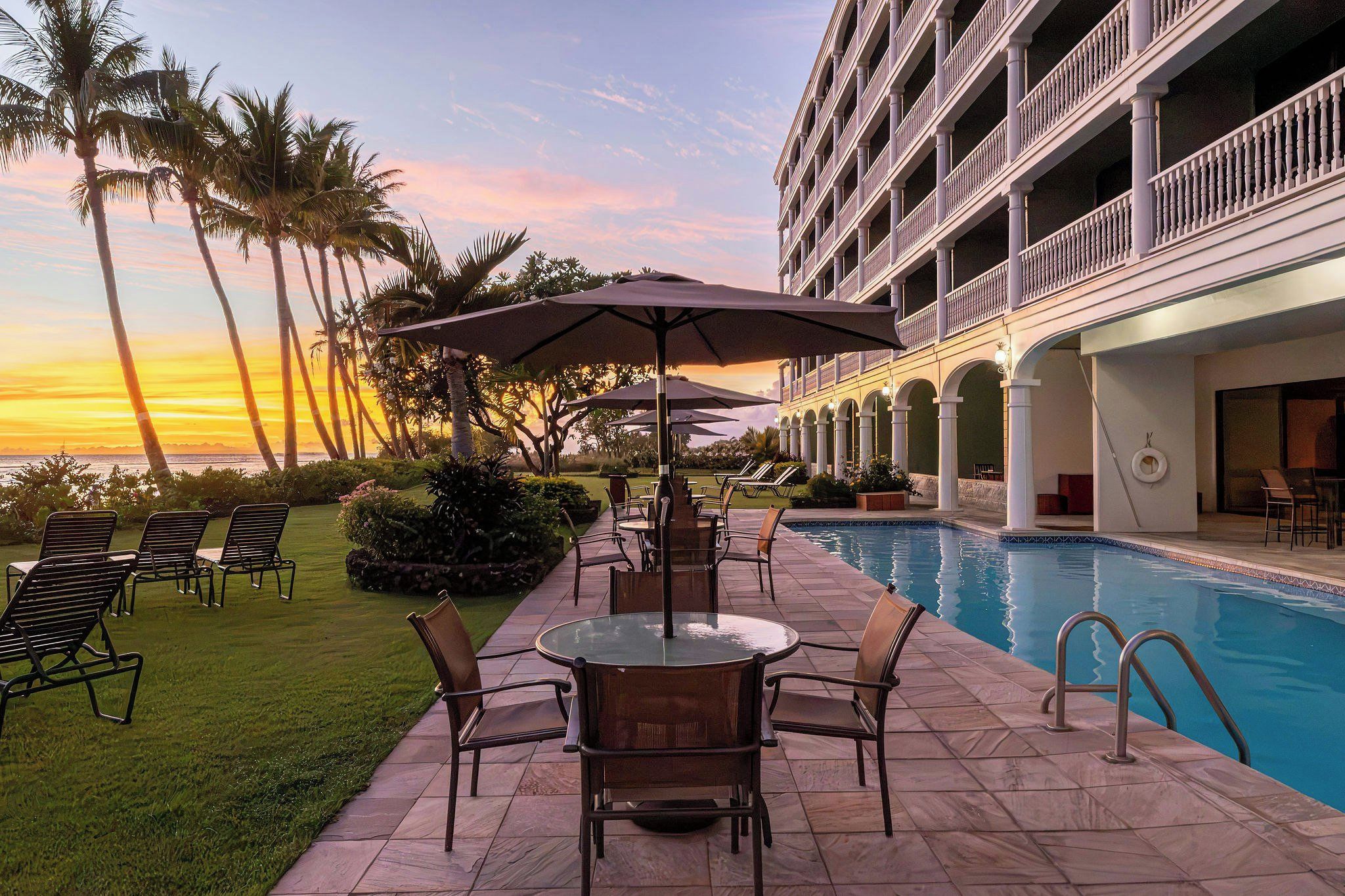 A poolside area with lounge chairs, a table, and umbrellas, surrounded by palm trees and a building with balconies.