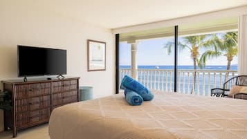 A hotel room with a bed, a television on a wooden dresser, and a balcony with a view of the ocean and palm trees.