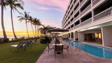 A hotel pool area with lounge chairs and umbrellas, surrounded by palm trees and a view of the ocean.