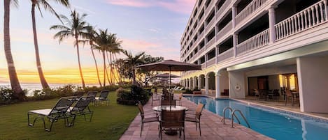 A hotel pool area with lounge chairs and umbrellas, surrounded by palm trees and a view of the ocean.