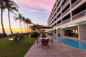 Un espace piscine d’hôtel avec des chaises longues et des parasols, entouré de palmiers et offrant une vue sur l’océan.