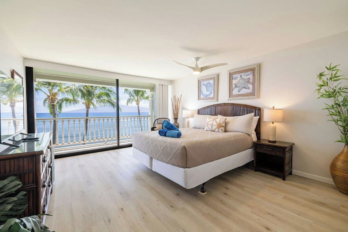 Un dormitorio con cama, mesita de noche, lámpara, ventana grande con vistas a palmeras y al mar, y ventilador de techo.