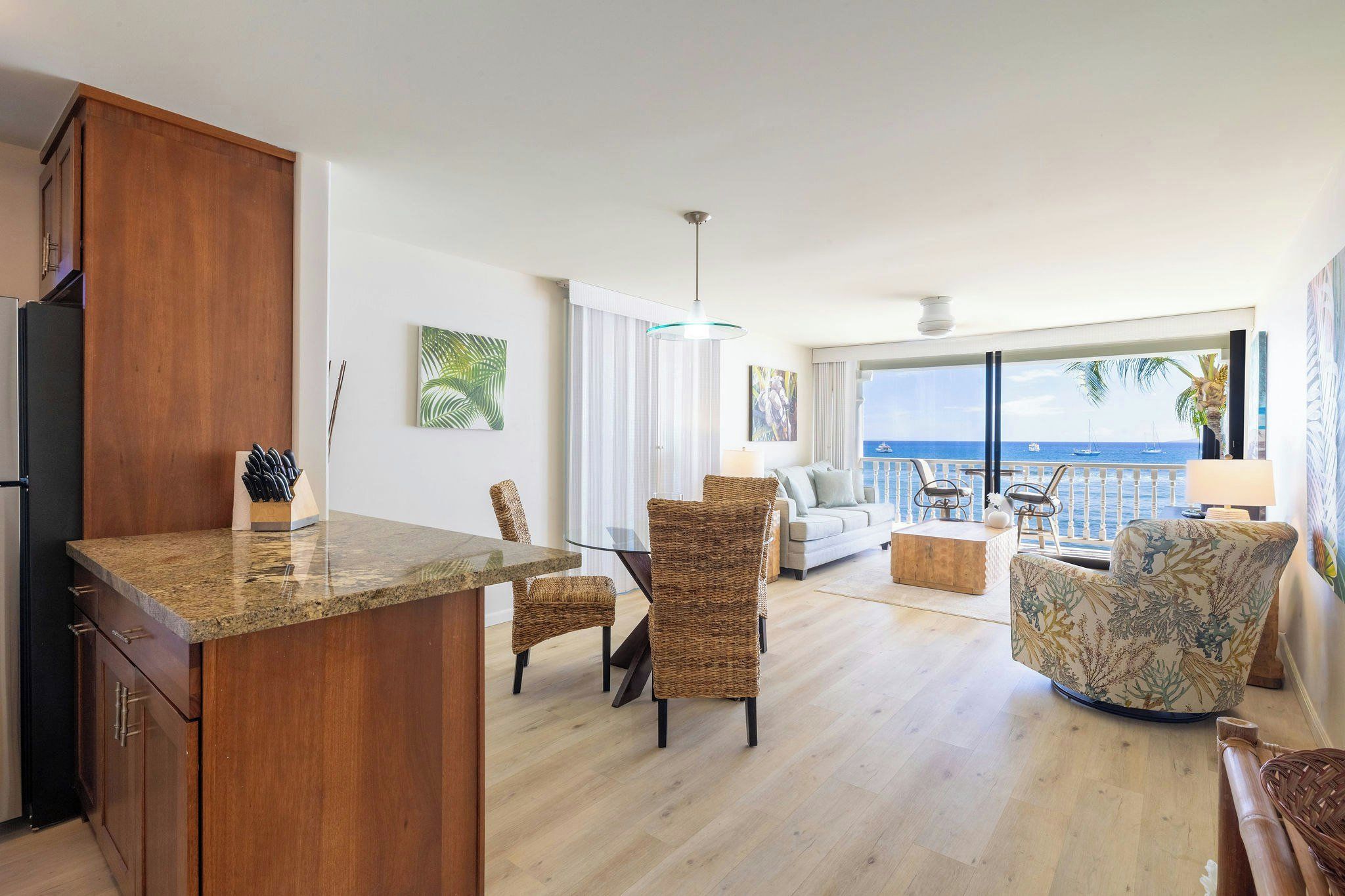 A modern living room with a kitchen area, featuring a large window with a view of the sea, wicker furniture, and a wooden cabinet.
