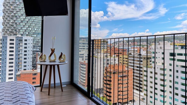 Ein modernes Hotelzimmer mit großem Fenster, einem Flachbildfernseher, einem runden Holztisch mit Dekoration und einem Bett mit gestreiftem Überwurf.