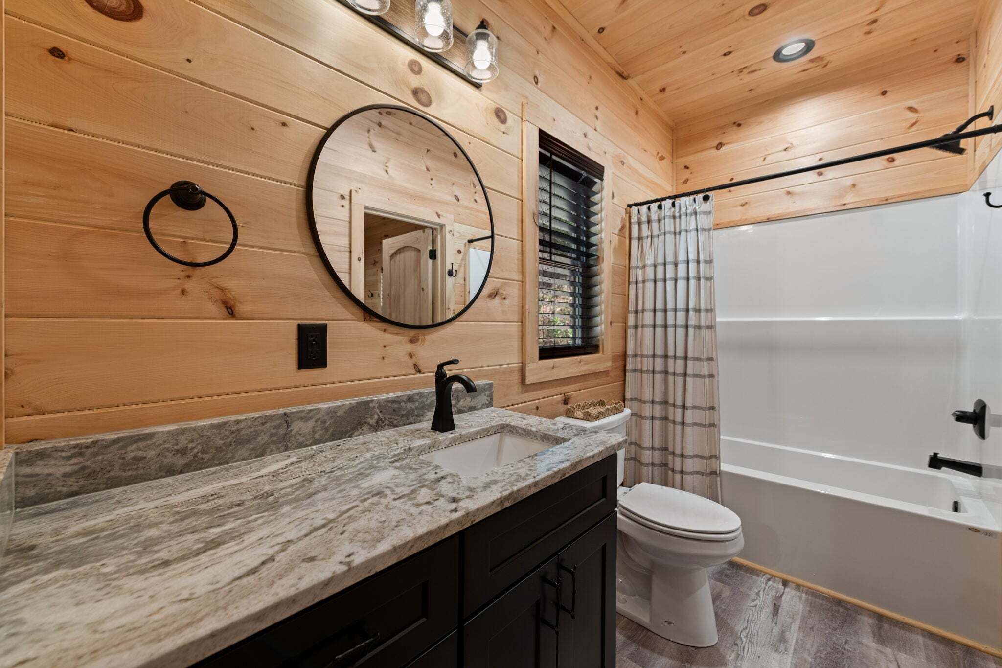 A wooden interior bathroom with a marble countertop, a round mirror, a toilet, and a bathtub.