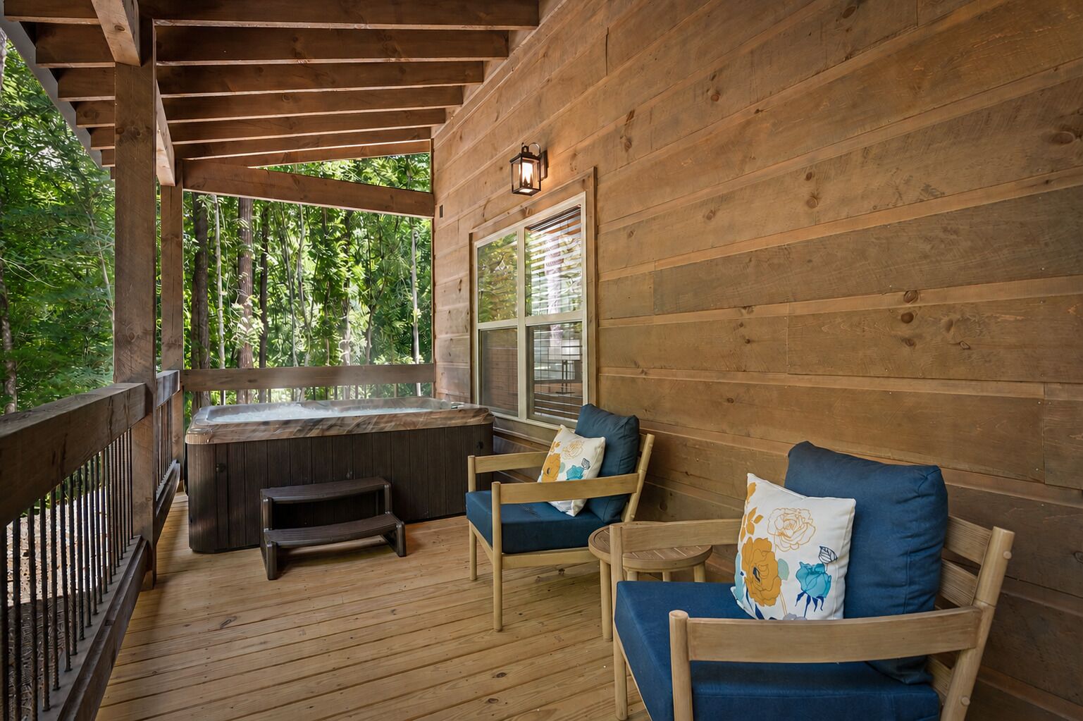 A wooden deck with a hot tub, two chairs with cushions, and a window overlooking greenery.