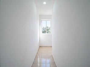 Un pasillo estrecho con paredes blancas y piso de baldosas, que lleva a una ventana con vistas a árboles.