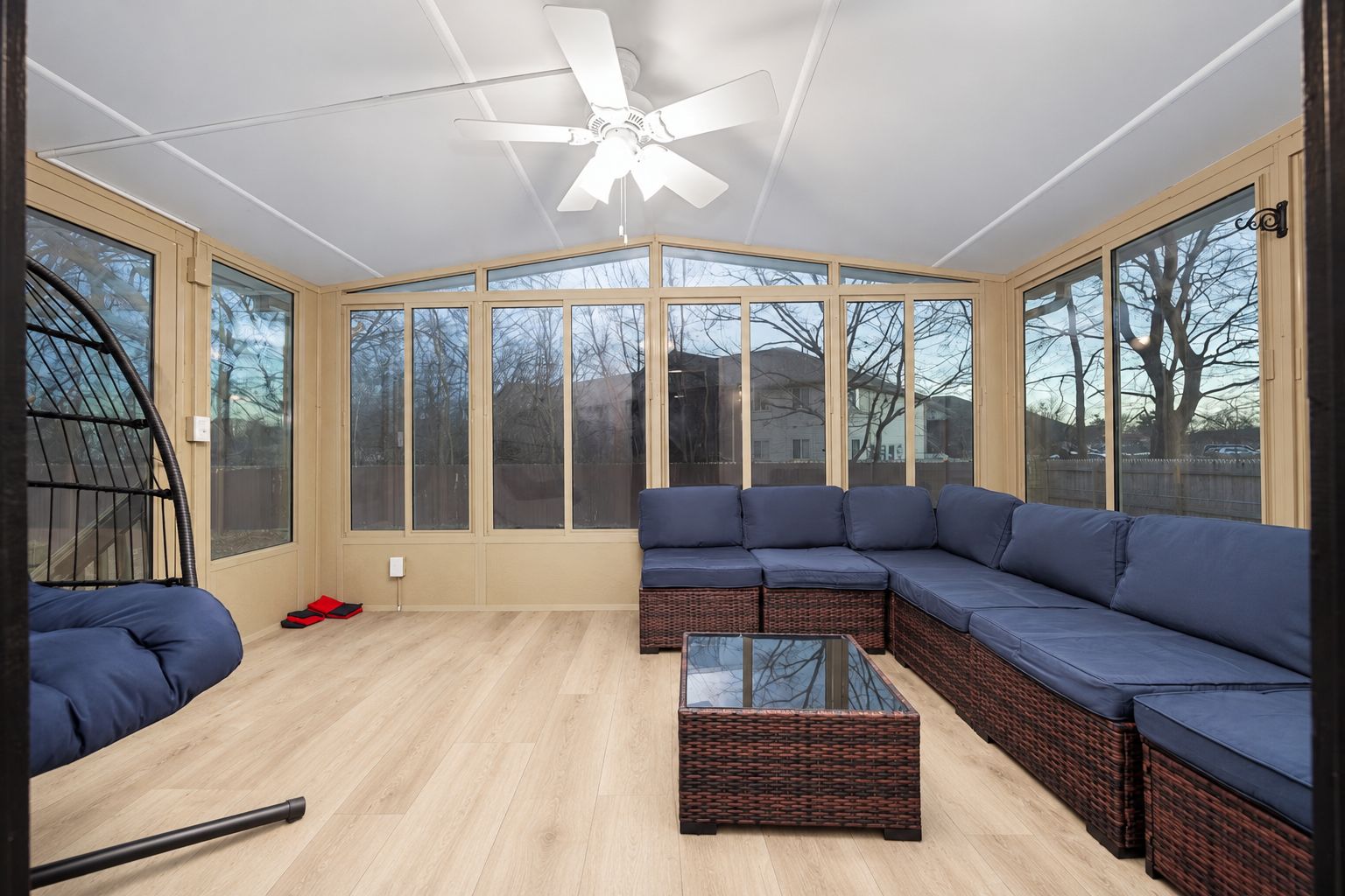 A sunroom with a large glass enclosure, a ceiling fan, a sectional sofa, and a glass coffee table.