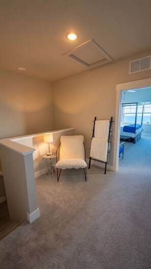 A room with a white chair, a small table with a lamp, a ceiling vent, and a bed with a blue bedspread.