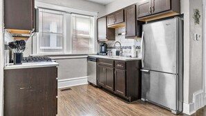 A kitchen with dark wooden cabinets, a stainless steel refrigerator, a dishwasher, and a window with blinds. - The Oak | Pet-Friendly King Retreat (Saint Louis)
