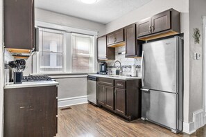 A kitchen with dark wooden cabinets, a stainless steel refrigerator, a dishwasher, and a window with blinds. - The Oak | Pet-Friendly King Retreat (Saint Louis)