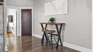 A hallway with a wooden floor, a dark door, a white wall with a framed artwork, and a small table with stools. - The Oak | Pet-Friendly King Retreat (Saint Louis)