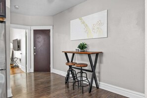 A hallway with a wooden floor, a dark door, a white wall with a framed artwork, and a small table with stools. - The Oak | Pet-Friendly King Retreat (Saint Louis)