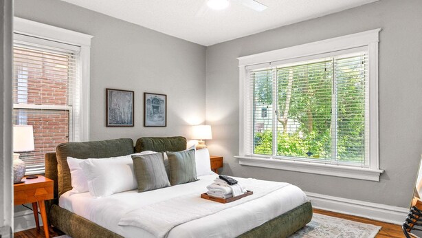 A bedroom with a bed, a nightstand, a lamp, a window with blinds, and a view of greenery outside. - The Oak | Pet-Friendly King Retreat (Saint Louis)