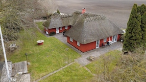 En lantlig fastighet med två stugor med halmtak, en gräsklädd innergård och ett stort, öppet fält i bakgrunden.