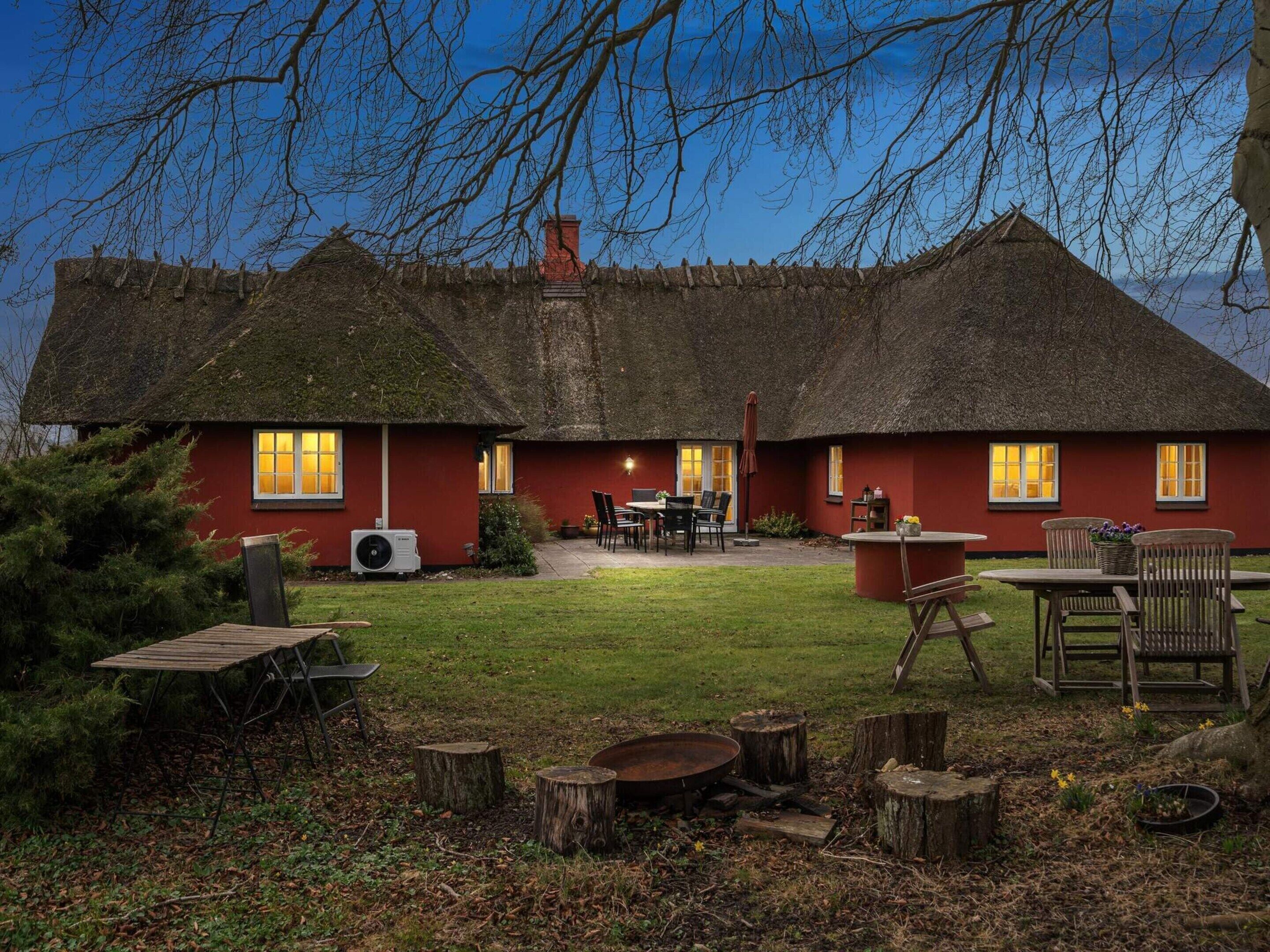 A thatched-roof cottage with a red exterior, a patio with outdoor seating, and a well-maintained lawn.