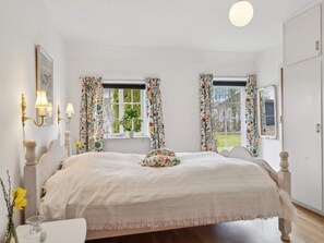 Ein Schlafzimmer mit einem Bett, zwei Fenstern mit Blumenvorhängen, einem Nachttisch und einem Beistelltisch.