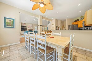 Ein Essbereich mit Holztisch und weißen Stühlen, Deckenventilator und einer Küche im Hintergrund.