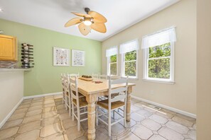 A dining area with a hexagonal tile floor, a wooden table, white chairs, a ceiling fan, and a window with white valance. - Tuck Away: New Rental, Dog-friendly, Private Pool (Corolla)