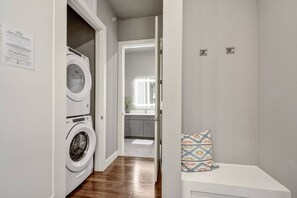 A laundry room with stacked washers and dryers, a bench with a patterned cushion, and a door leading to a kitchen area. - Modern Luxury 1BR: Free Parking + Fast WiFi (San Antonio)