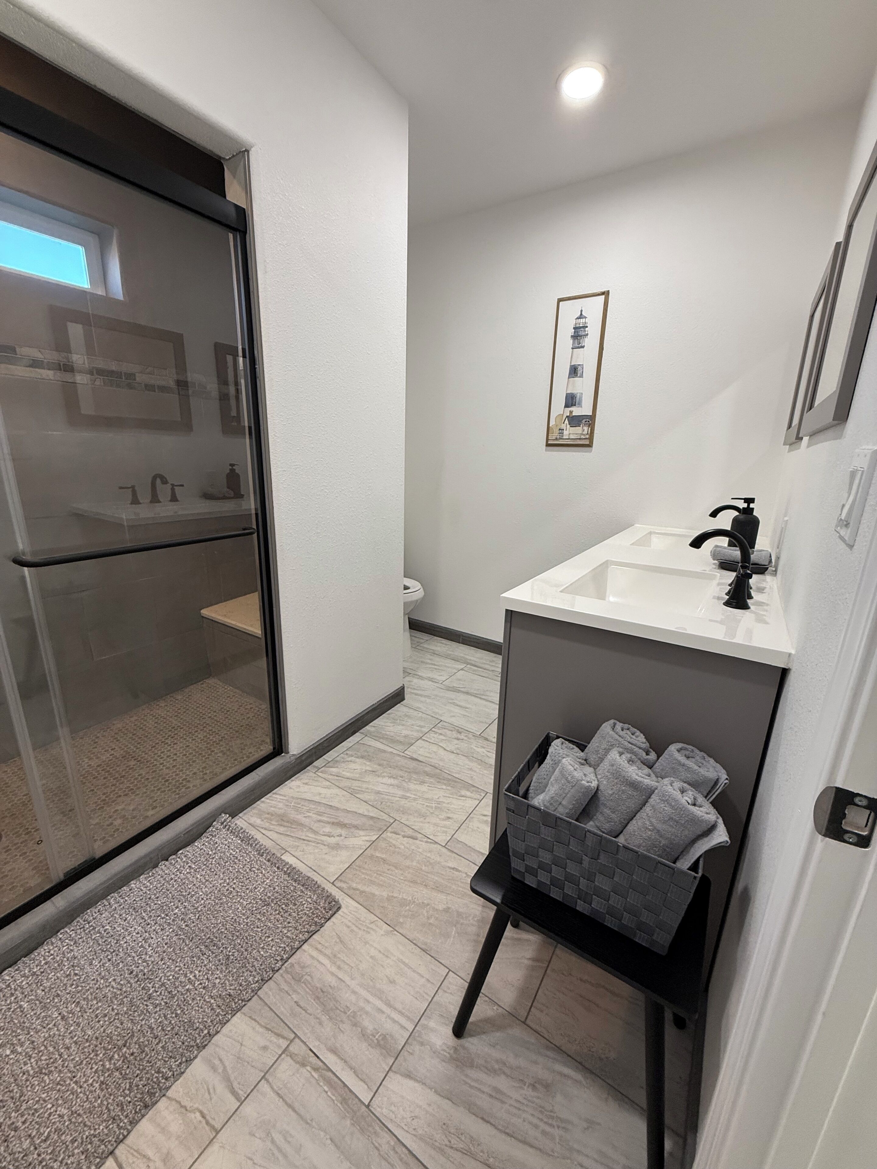 Un baño moderno con cabina de ducha de vidrio, un lavamanos blanco con grifo negro, un cuadro en la pared y un perchero con toallas.