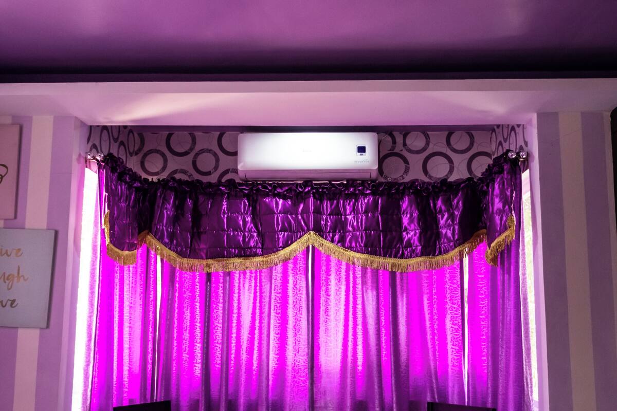 Ein Zimmer mit einem Bett, einem Sessel, einem kleinen Tisch, violetten Vorhängen und einer Klimaanlage an der Wand.
