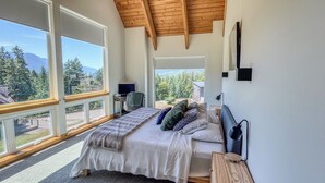 A bedroom with a large bed, a desk, a chair, and a TV. There are large windows with a view of trees and mountains. - The Lookout | Wifi, AC, Modern (Packwood)