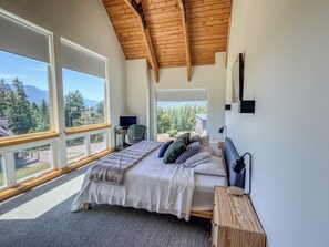 Ein Schlafzimmer mit einem großen Bett, einem Schreibtisch, einem Sessel und einem Fernseher. Große Fenster bieten Blick auf Bäume und Berge.