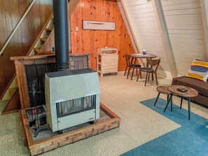 A cozy room with a wood-burning stove, a dining area with chairs and a table, and a staircase leading to an upper level. - Forest Echo A-Frame | Wifi, Hot Tub (Packwood)