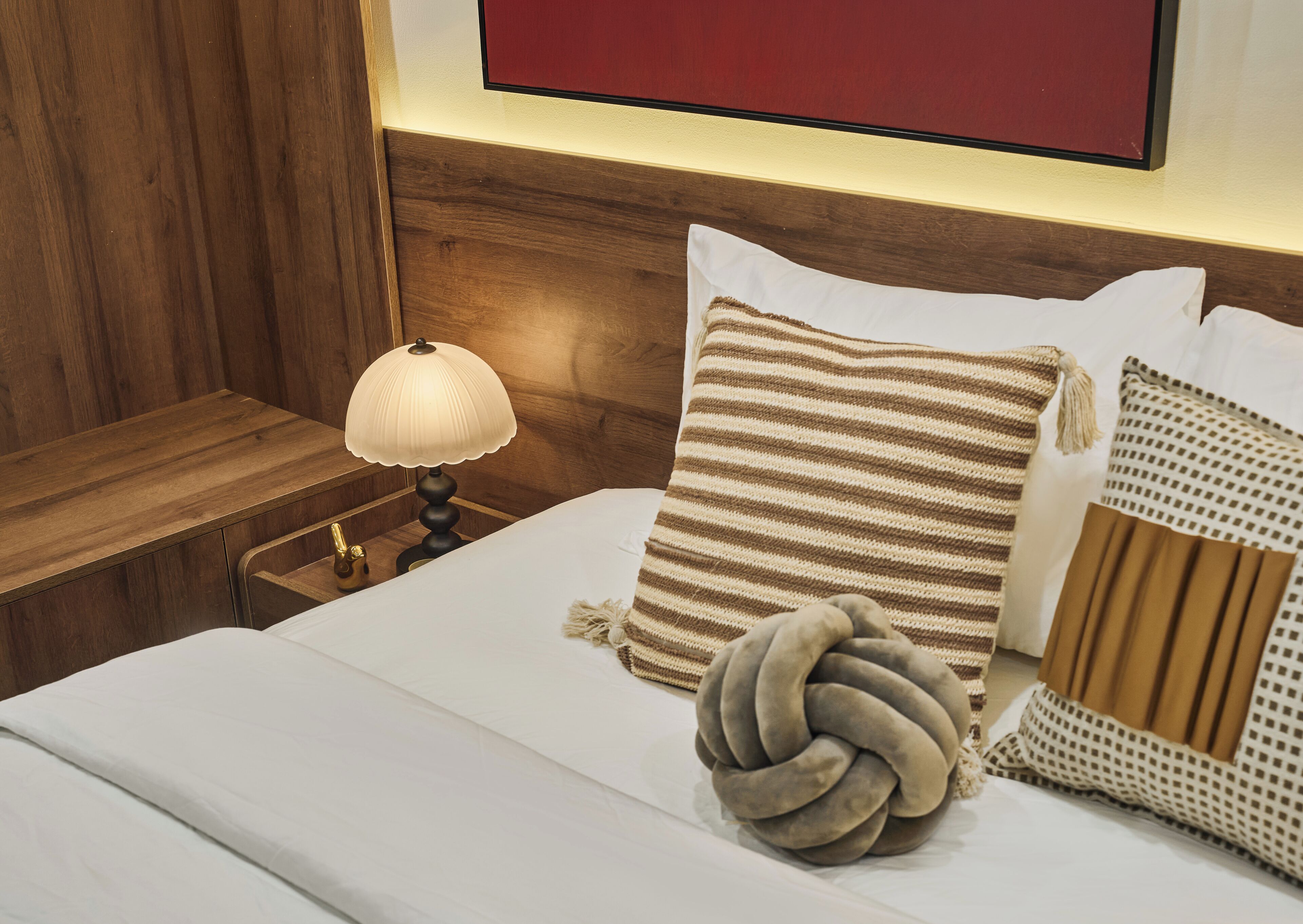Una cama bien tendida con un cojín a rayas, un cojín decorativo redondo y una lámpara en una mesita de noche.